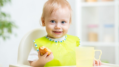 Criança de babete amarelo sentada numa cadeira de refeição para bebés com comida numa mão e um recipiente de plástico à frente.